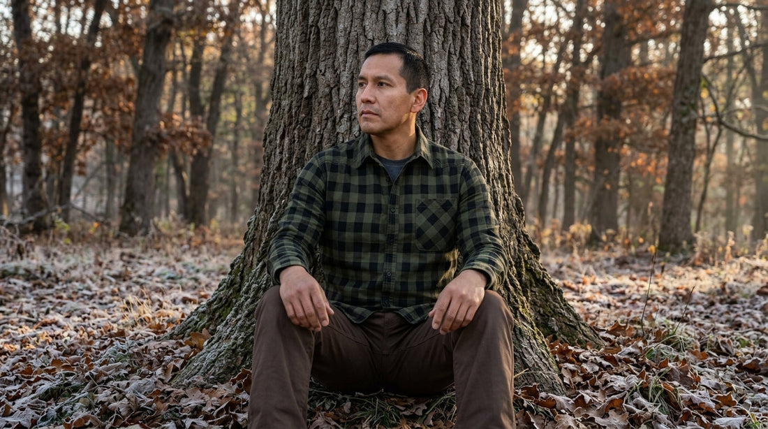Hunter in a green buffalo-check flannel shirt sitting quietly against an oak in a frosty Wisconsin forest at dawn
