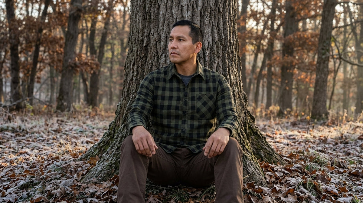 Hunter in a green buffalo-check flannel shirt sitting quietly against an oak in a frosty Wisconsin forest at dawn
