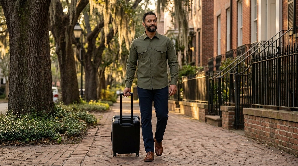 Man in army green utility shirt and navy chinos pulling a carry-on down a Savannah brick sidewalk, business casual travel shirt